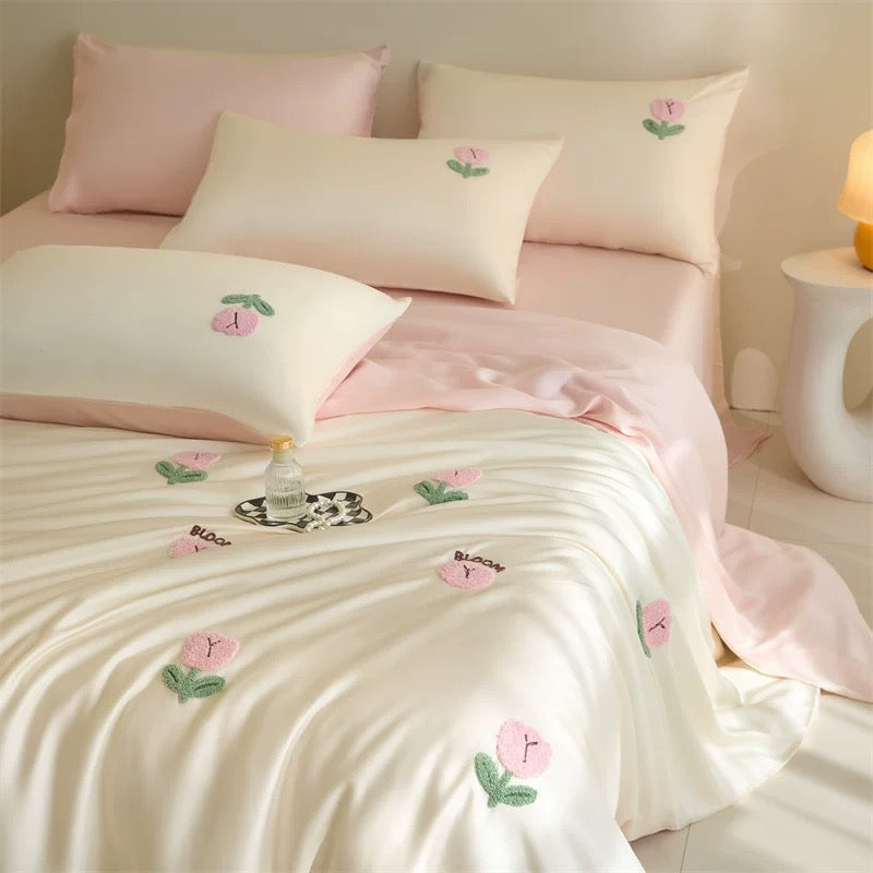 A neatly made bed with a soft cream bedding set and pale pink sheets, decorated with embroidered pink flowers and green leaves. Several matching pillows are arranged on the bed, with a small decorative tray and a lit lamp visible in a softly lit bedroom setting.
