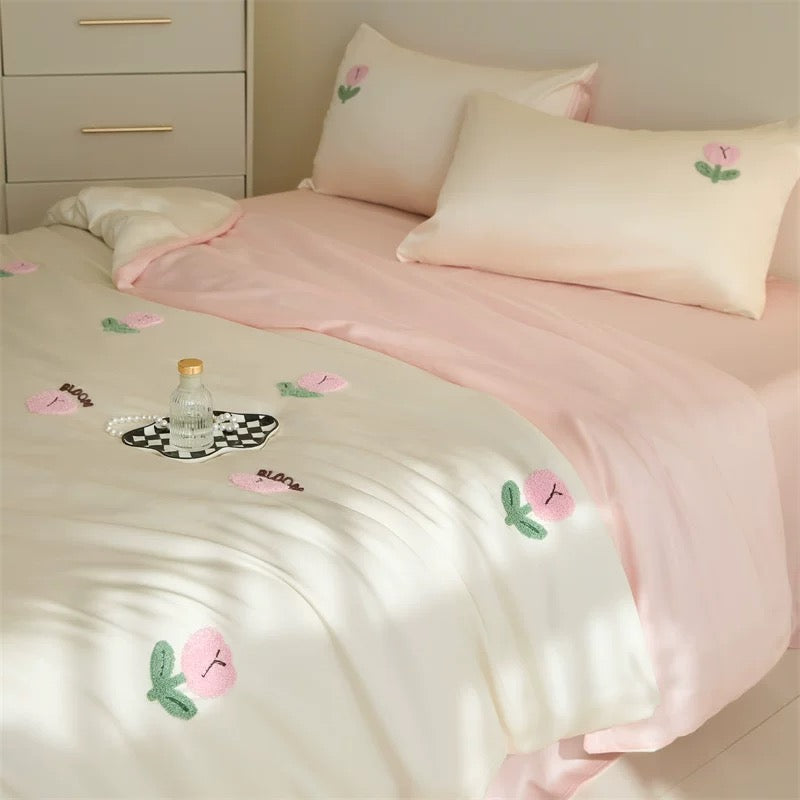 A neatly made bed with a soft pink and off-white bedding set featuring embroidered peach motifs. The scene is styled in a bedroom with a light wood dresser in the background.