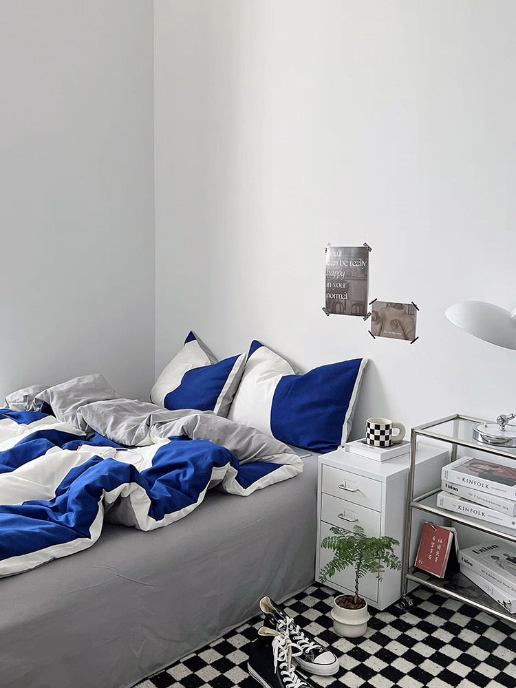 Bedroom with gray bedding and blue pillows, nightstand with books, and checkered floor.