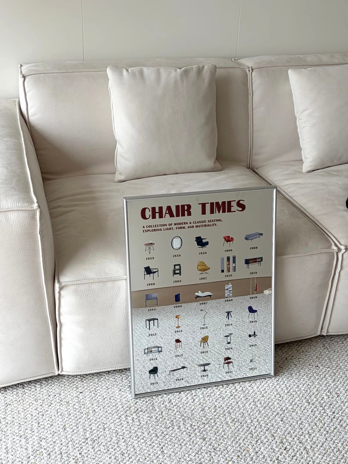 A framed 'CHAIR TIMES' furniture reference chart leaning against a cream-colored leather sofa on a textured light gray carpet.