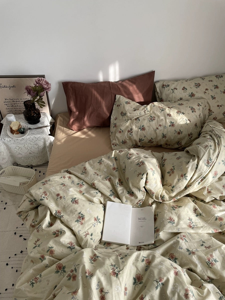 Vintage floral duvet on a sunlit bed with rust pillows, open book, flowers, and bedside table.
