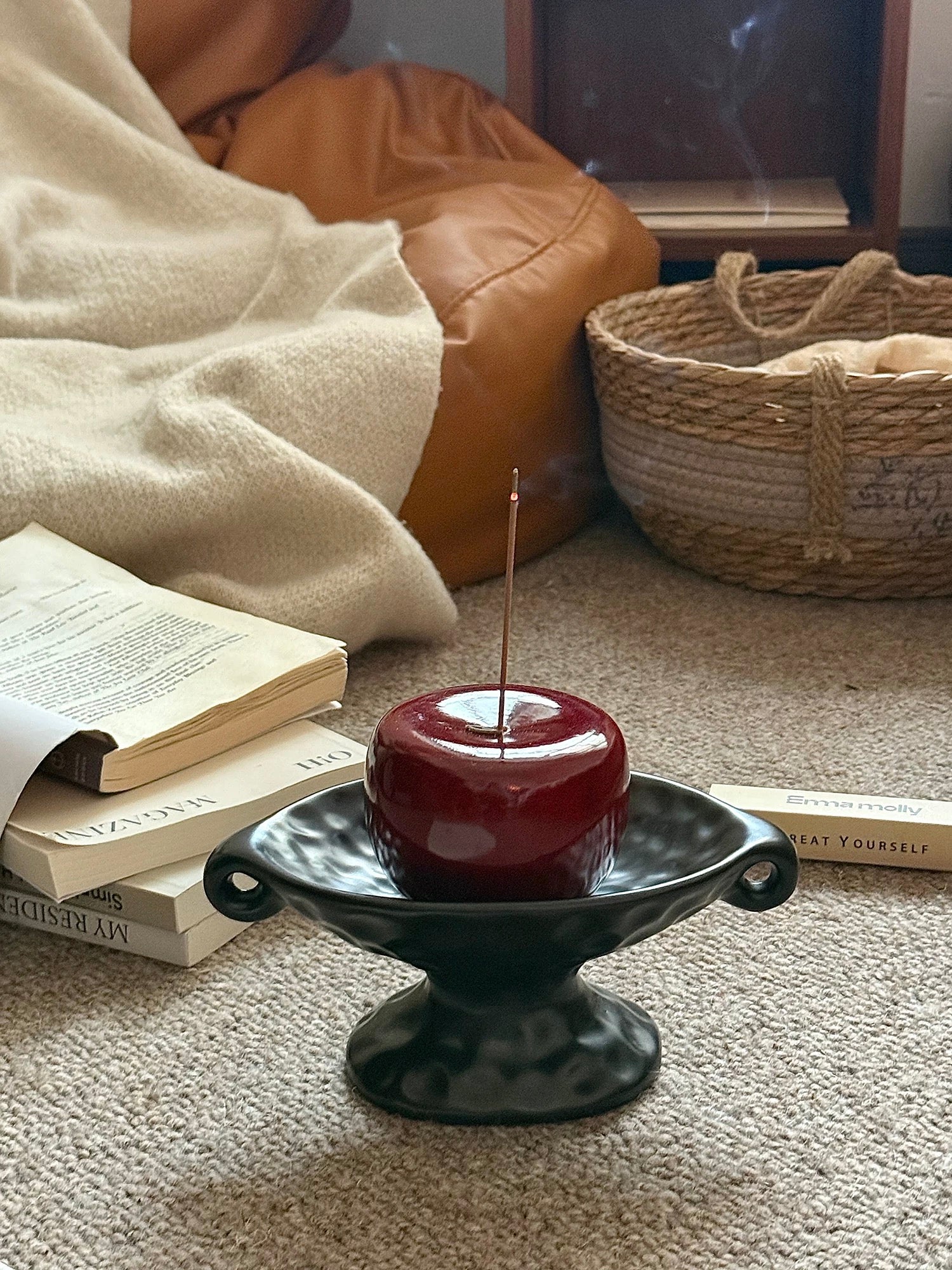 A dark red apple-shaped candle in a black pedestal holder, placed on a carpeted floor in a cozy living room setting with books, a blanket, and a woven basket in the background.