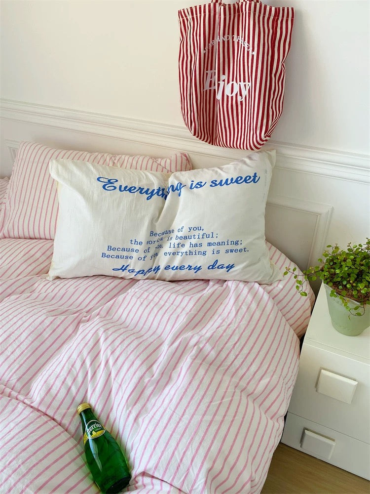 Bed with pink striped bedding, white pillow with blue text, and red and white striped bag on a white wall.