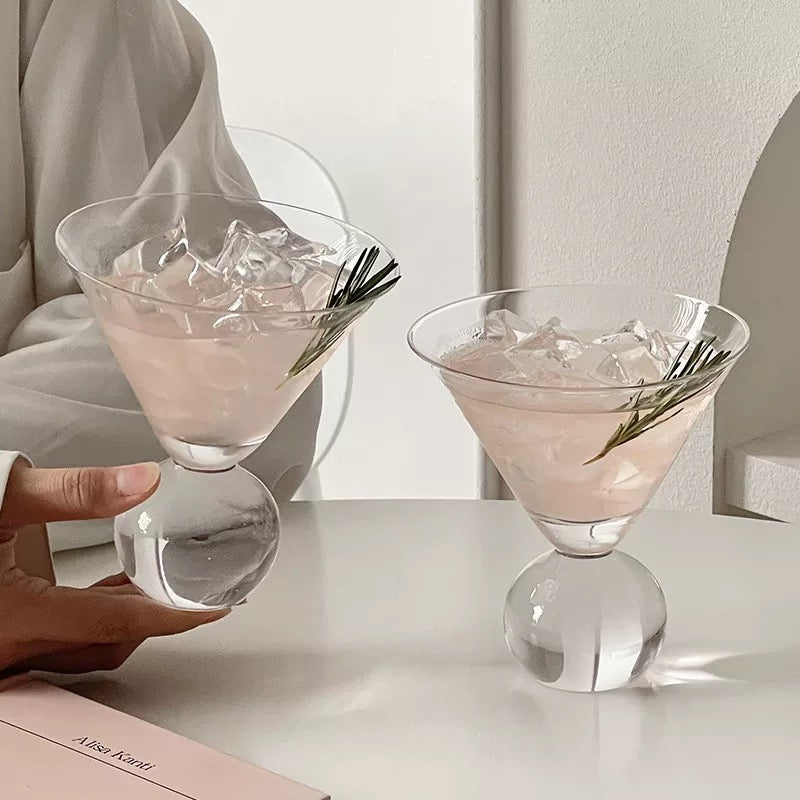 Two pink cocktails with ice and a sprig of rosemary in clear glasses on a white surface.