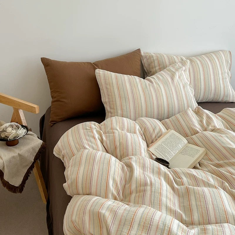 A cozy bed styled with a cream-colored cotton duvet cover and matching pillowcases featuring thin vertical stripes in muted red, orange, and green, accented by solid brown pillows and a brown bed frame.
