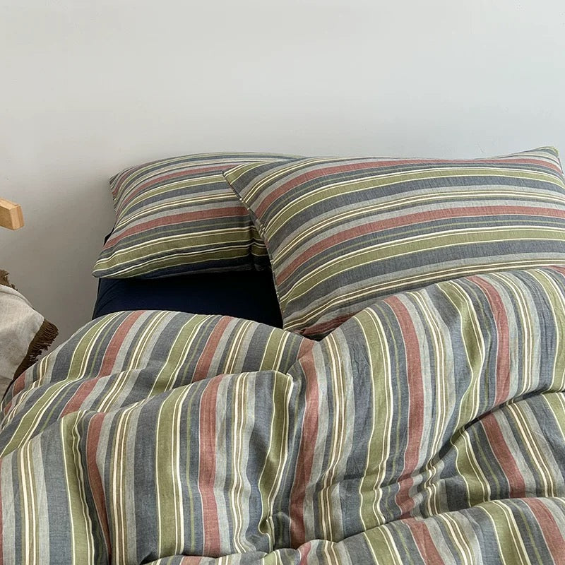 A close-up of a striped bedding set with matching pillowcases, featuring vertical stripes in muted gray, olive green, dusty red, and cream, styled on a bed against a plain light gray wall.