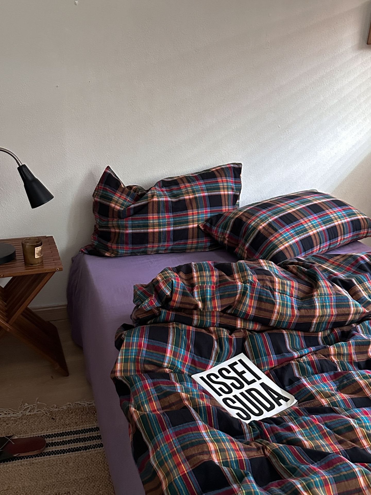 A bed in a bedroom styled with a multicolor plaid bedding set, featuring a purple sheet underneath. The plaid pattern contains shades of red, green, blue, and black.