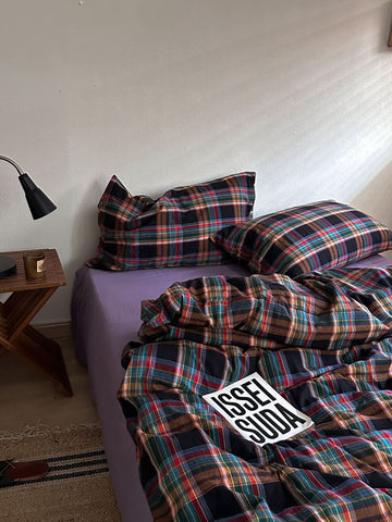 A bed in a bedroom styled with a multicolor plaid bedding set, featuring a purple sheet underneath. The plaid pattern contains shades of red, green, blue, and black.