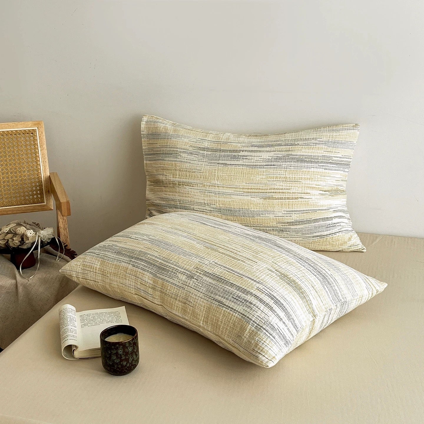 Two rectangular throw pillows with a textured, horizontal striped pattern in beige, cream, and gray, styled on a beige sofa next to a woven chair and a candle.