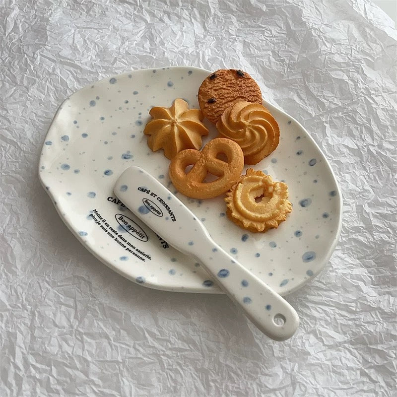 A white ceramic plate with blue speckles and a matching white spatula resting on it, styled with several golden-brown cookies on a crumpled white paper background.