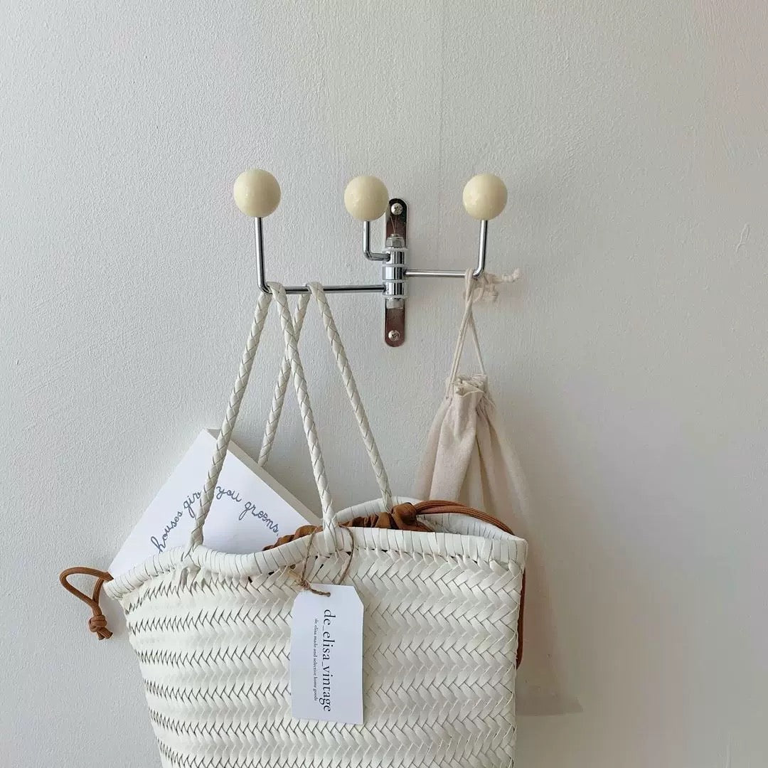 A cream woven tote bag with brown leather handles hangs from a silver wall hook with three cream spherical finials, against a plain off-white wall. A paper tag reading 'de luxe style' is attached to the bag.