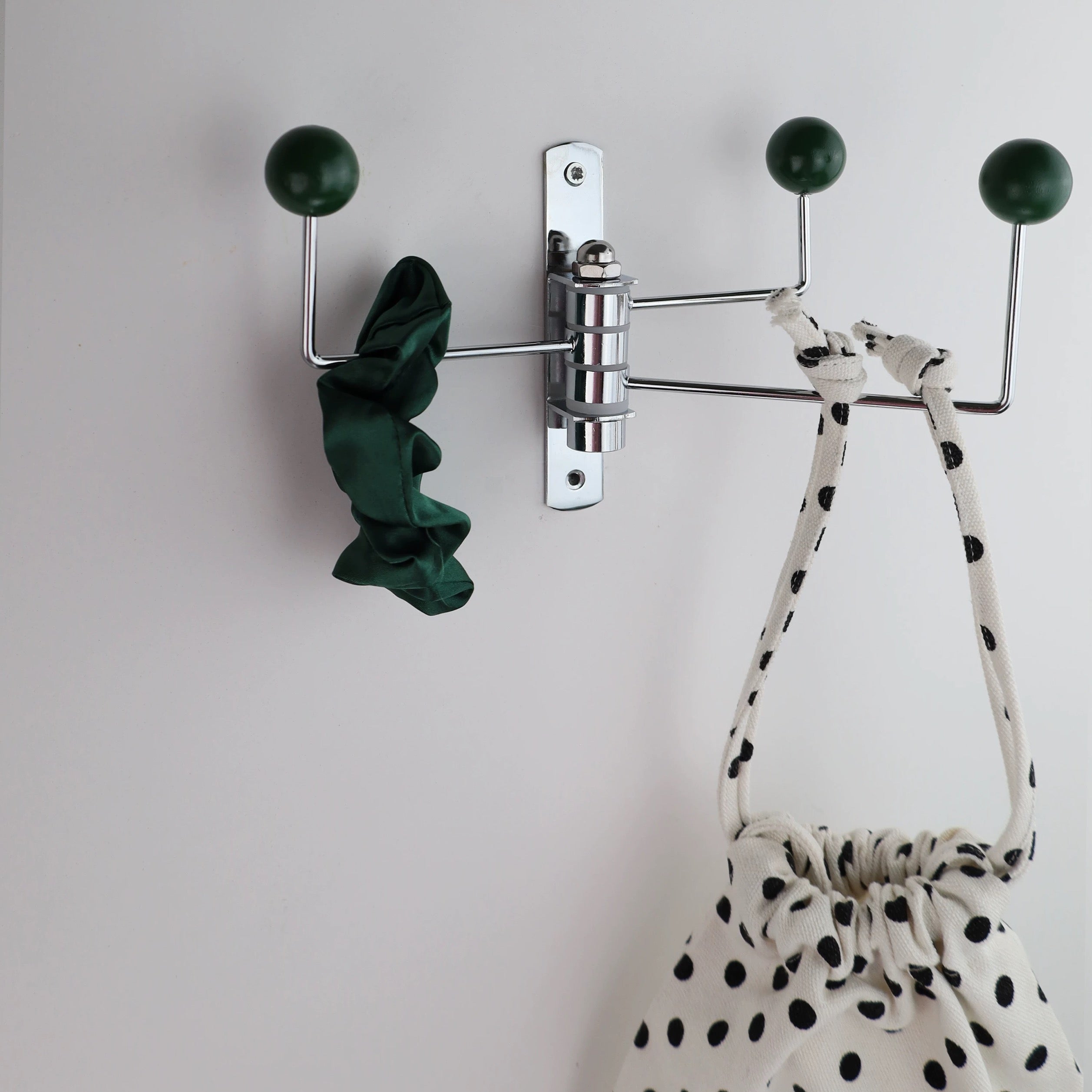 A wall-mounted three-arm coat rack with dark green spherical hooks and a polished silver metal frame, shown against a plain light wall with a dark green scrunchie and a black-and-white polka dot drawstring bag hanging from it.