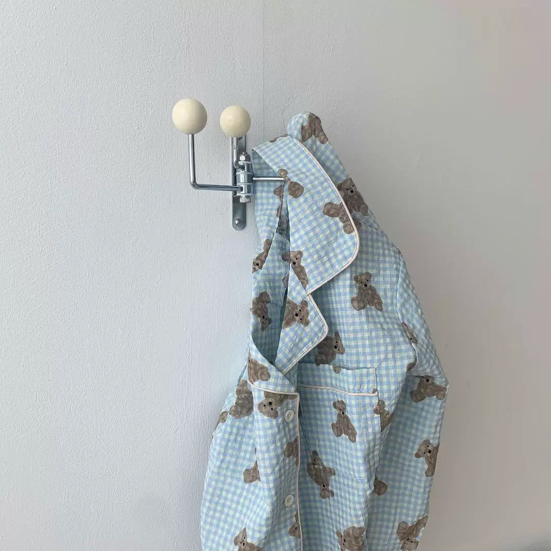 A light blue and white gingham pajama shirt with a brown teddy bear print hangs from a silver wall-mounted double hook with cream-colored spherical finials against a plain off-white wall.