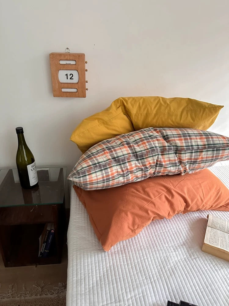 A mustard yellow pillowcase sits at the front of a bed with plaid and brown designs behind it.
