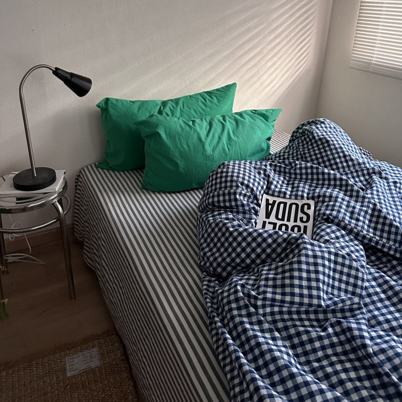 A bed in a real bedroom setting with a blue-and-white gingham duvet, striped bedding, two green pillows, a black floor lamp on a metal side table, and a window with blinds casting diagonal shadows.