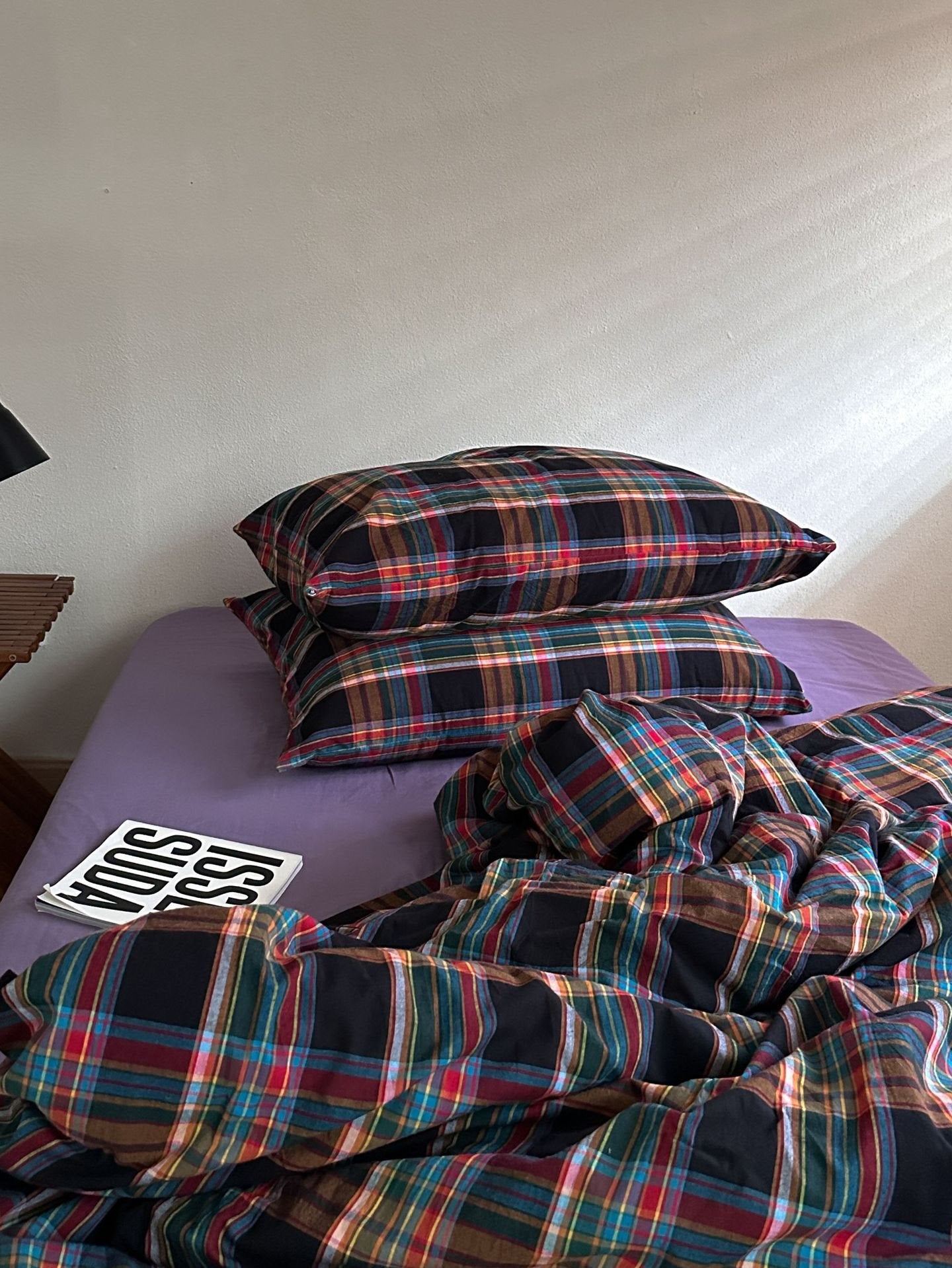 A bed with a purple sheet and a multicolor plaid bedding set, including two pillows and a rumpled duvet cover, in a bedroom setting.