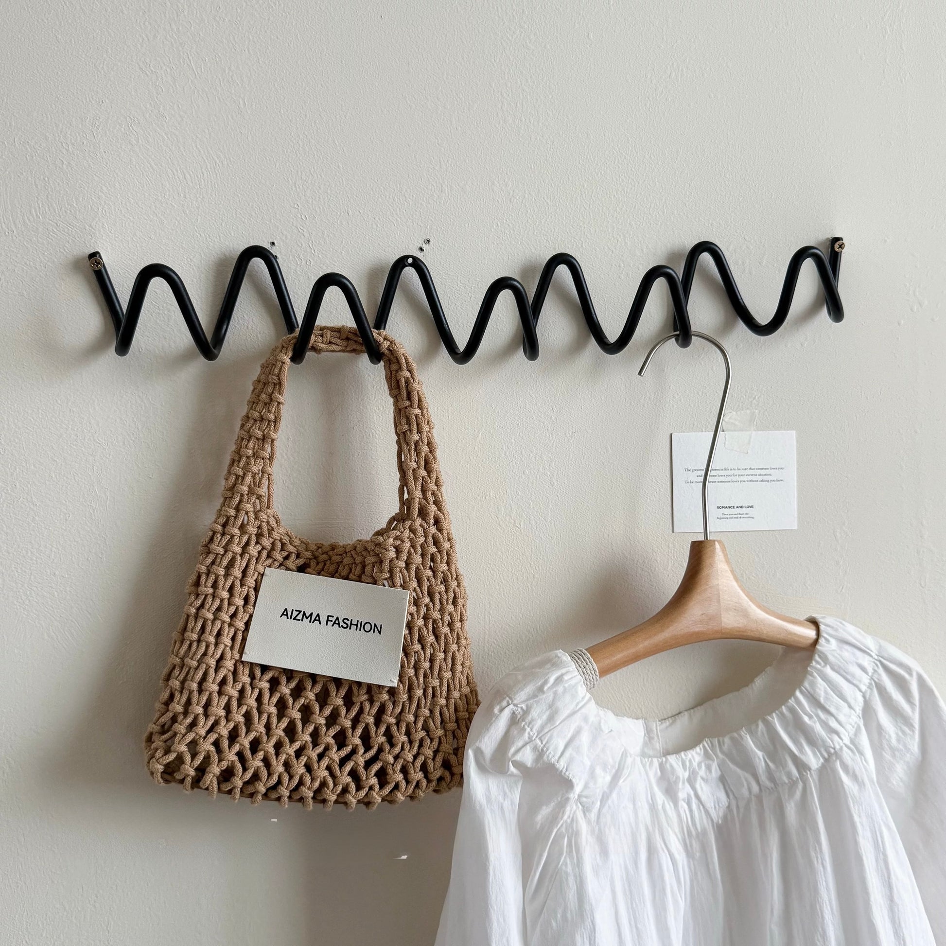 Wavy black metal hook with a woven bag and white garment on a light gray wall.