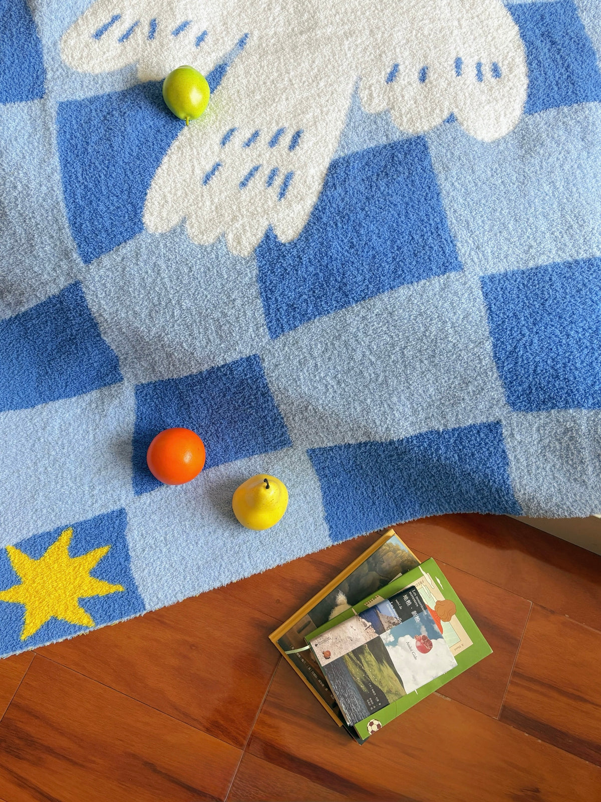 A blue and white checkered plush rug with a white dove motif and a yellow star, styled on a wooden floor with small colorful toy fruits and a stack of children's books.