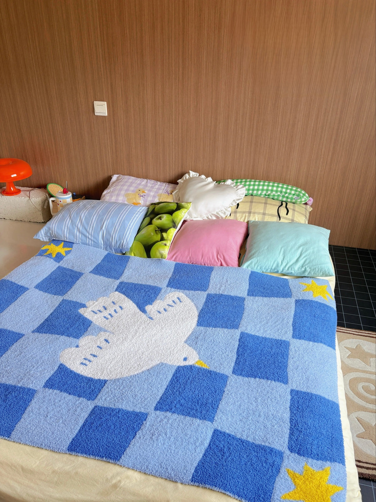 A blue checkered fleece blanket with a white dove design and yellow star accents spread across a bed, surrounded by various decorative pillows in a bedroom setting.