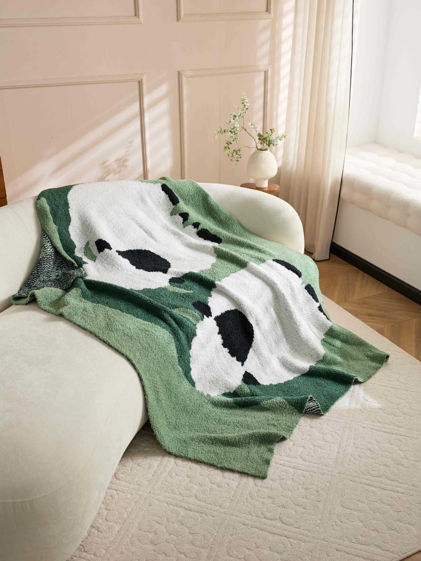 A green and white knit blanket with a black and white panda print is draped over a cream-colored sofa in a softly lit room with a window and curtains.