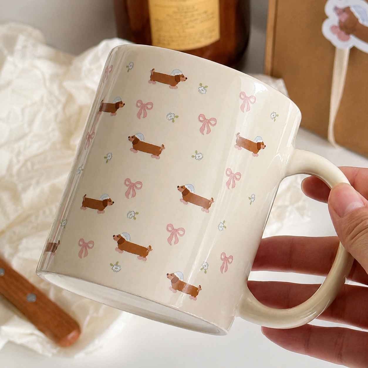 A hand holding a cream-colored ceramic mug decorated with brown dachshund illustrations, pink bows, and small white flowers, with a soft, styled background.