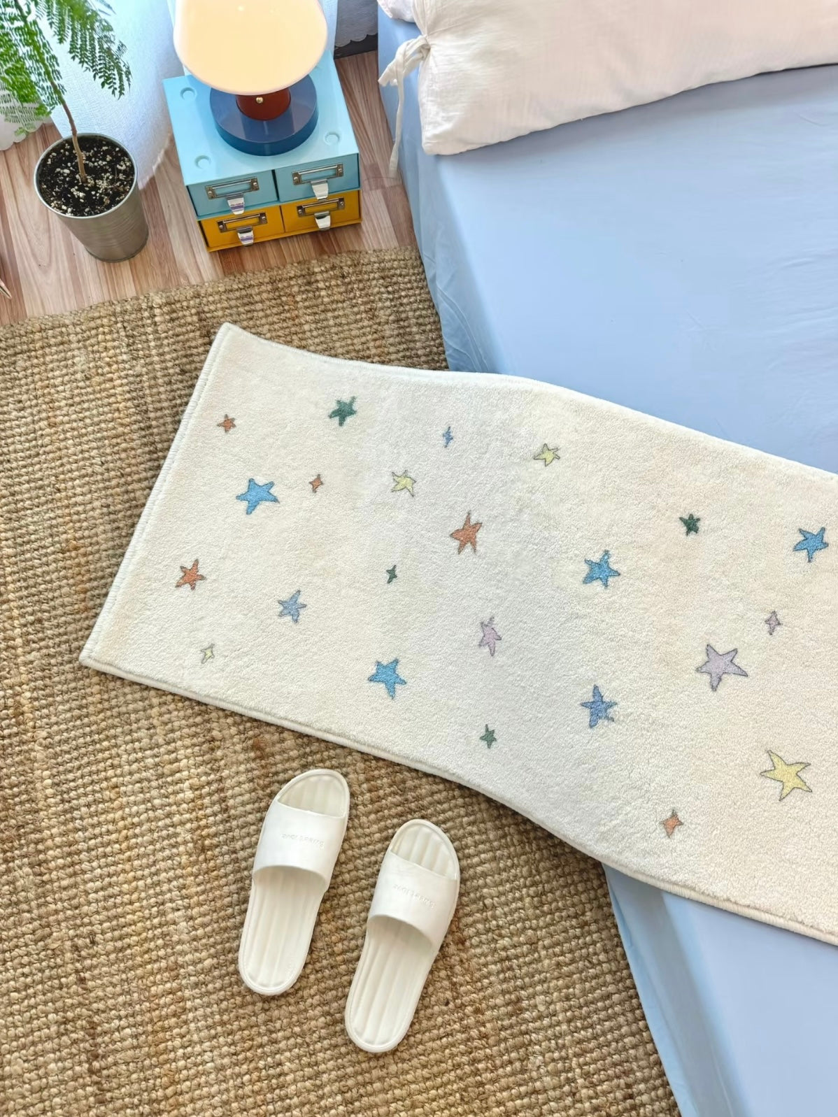 A Y2K rug with multicolor stars on a cream background lies diagonally beside a bed and white slippers.