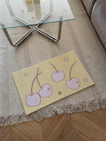 A rectangular beige rug with a fringed edge lies on a wooden floor in a living room setting. The rug features a whimsical pattern of three pink cherries with stems and small pink stars.