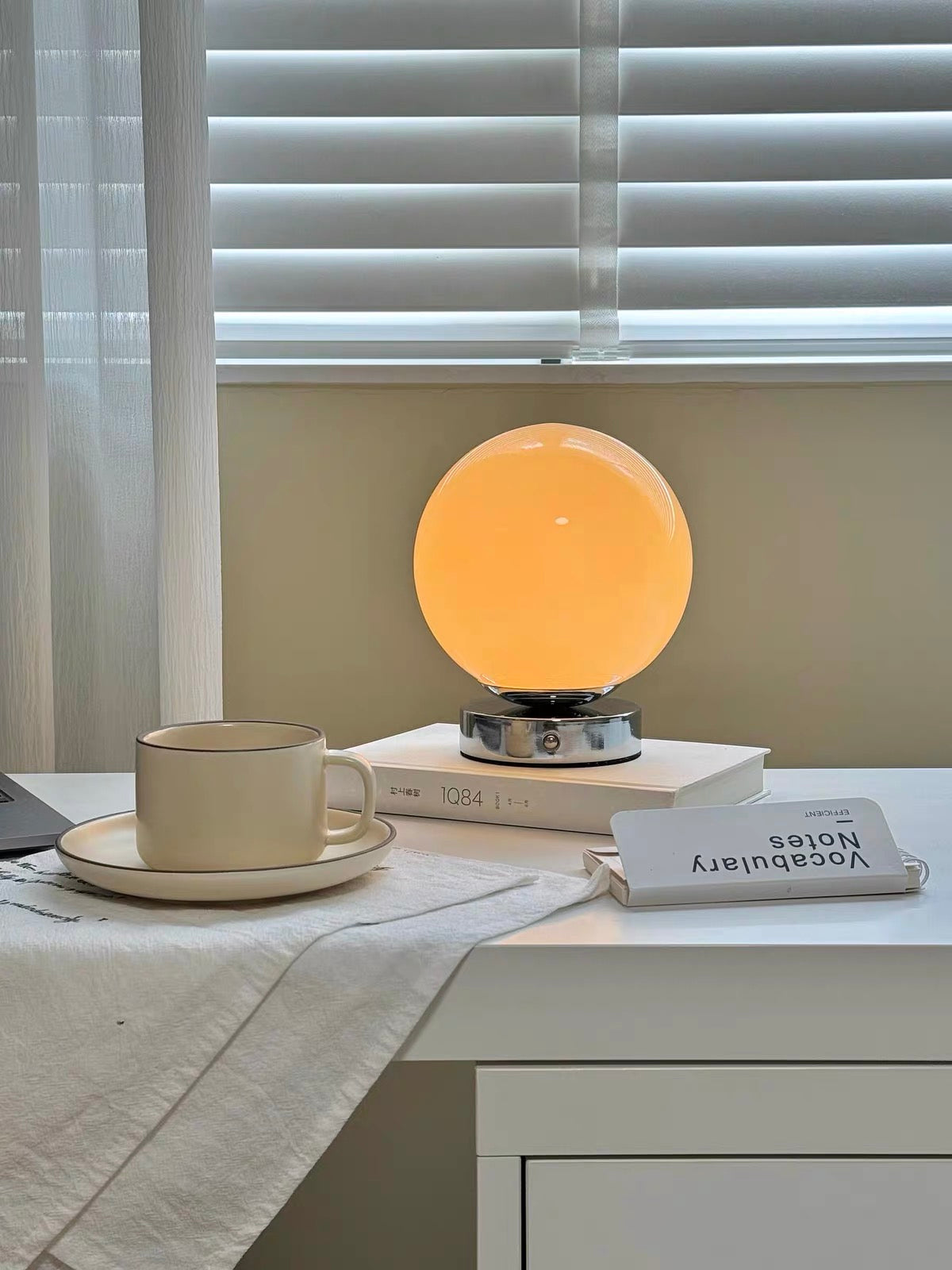 Table lamp with a round orange shade on a desk next to a cup and books, with blinds in the background.
