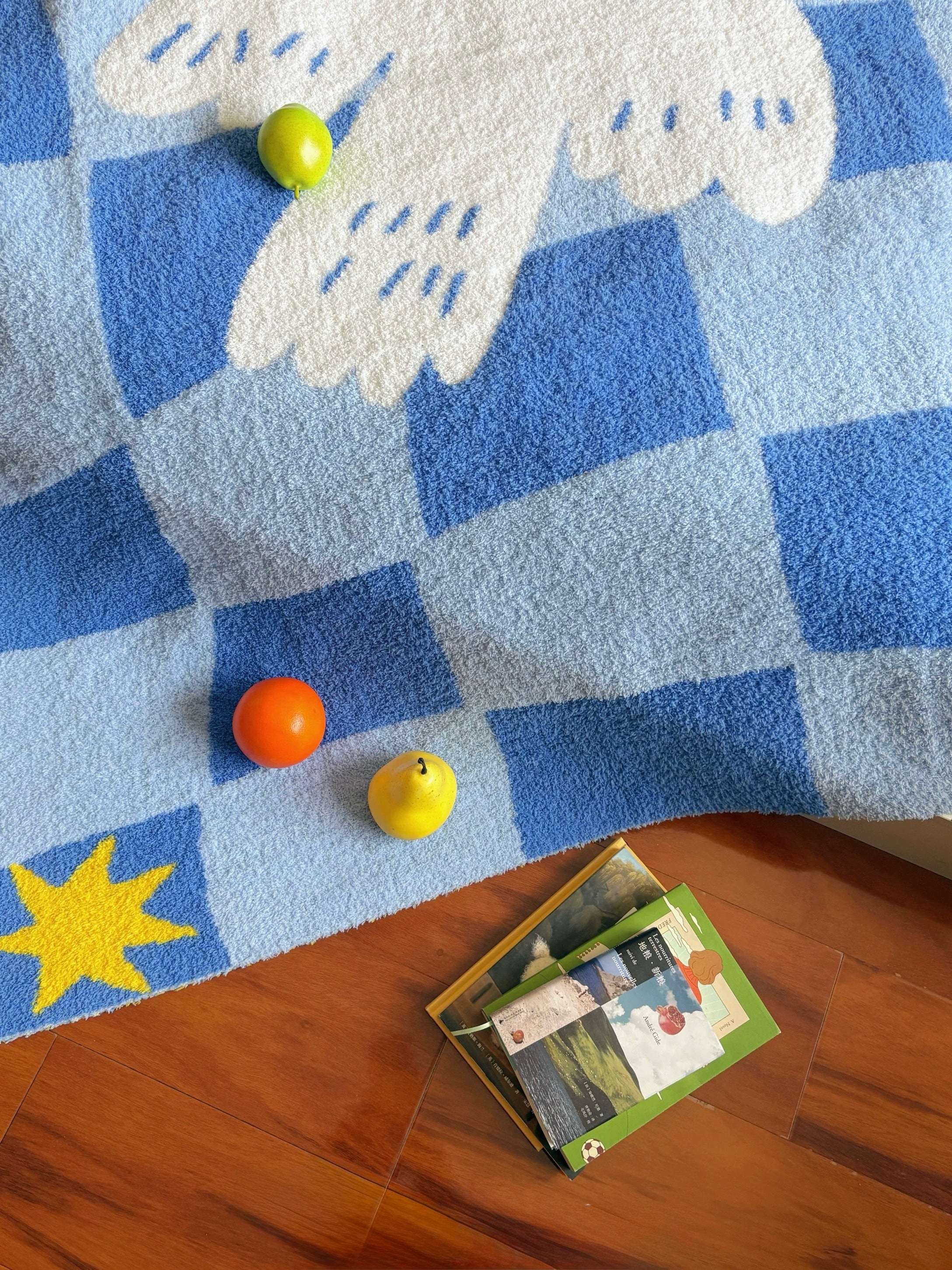 A blue and white checkered plush rug with a white dove motif and a yellow star, styled on a wooden floor with small toy fruits and a book.